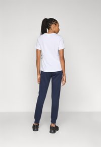 Vit t-shirt med kort ärm, kombinerad med marinblå sweatpants och svarta sneakers. Modellen har långa flätor stylade i en hästsvans. Minimalistisk design.