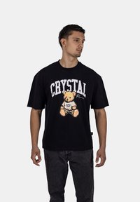Černé bavlněné tričko s grafikou medvídka a textem "CRYSTAL" v bílé barvě na jednobarevném pozadí.