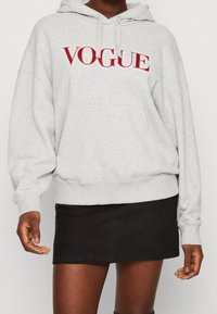 Felpa grigia con "VOGUE" ricamato in rosso sul petto, dotata di cappuccio con lacci e tasca frontale. Abbinata a una gonna nera.