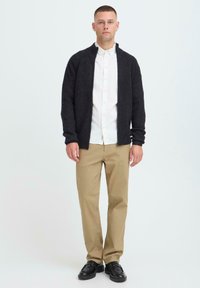 Donkere grijze cardigan, gebreide textuur, lange mouwen, open voorkant. Witte overhemden eronder, beige rechte broek, zwarte schoenen.