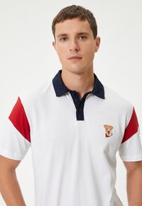 Polo skjorte i hvid med røde og marineblå accenter, der har en lille illustration af en hund på venstre bryst. Korte ærmer, krave-design.