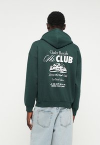 Zelená bavlnená mikina s kapucňou s grafickým potlačou na chrbte obsahujúcou "Chalet Royale Ski Club" s bielym textom a akcentmi.