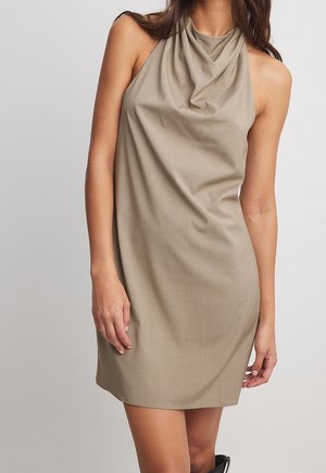 Kvinde iført en ærmeløs beige halterneck-kjole, stående mod en ensartet lys baggrund.