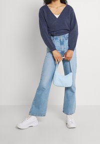 Femme portant un pull en V bleu marine, un jean bleu clair, des baskets blanches, tenant un petit sac à main bleu clair devant un arrière-plan uni.