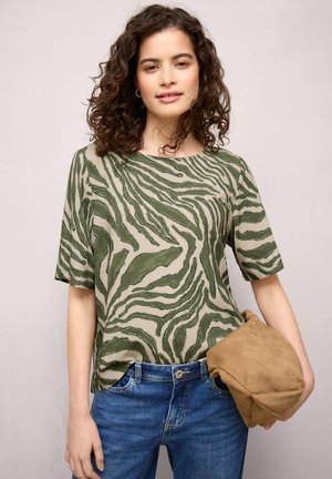 Ung kvinde med krøllet hår iført en grøn top med mønster og blå jeans, der holder en lys ruskindshat mod en ensfarvet baggrund.