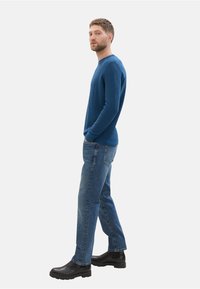 Suéter de punto azul, jeans azules, botas de tobillo negras. El suéter tiene un escote redondo y mangas largas. El tejido parece suave con una sutil textura.