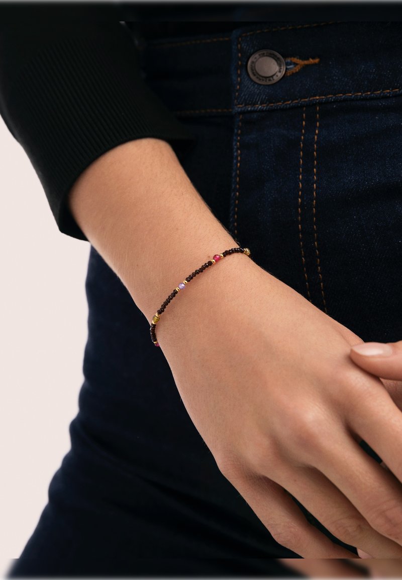 Zarter Armband mit kleinen, mehrfarbigen Perlen auf einem dünnen schwarzen Faden, getragen an einem Handgelenk vor einem dunklen Jeanshintergrund.