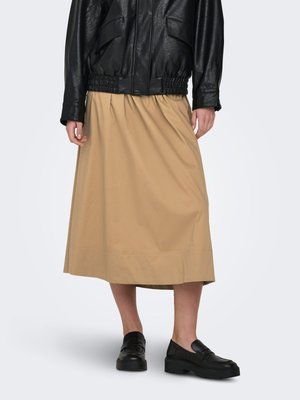 Vrouw die een beige rok tot op de knie, een zwart leren jasje en zwarte instapschoenen draagt, staand tegen een effen achtergrond.