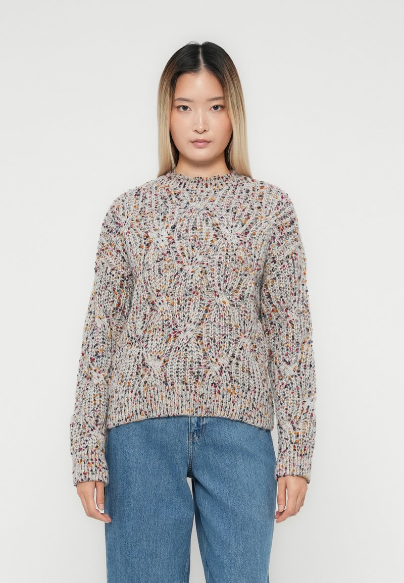 Vrouw in een veelkleurige gebreide trui met kabelpatronen en blauwe jeans, staand tegen een effen lichte achtergrond.