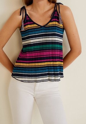 Kobieta w bezrękawniku w wielokolorowe paski z wiązanymi na ramionach ramiączkami oraz białych spodniach, stojąca na tle beżowej ściany.