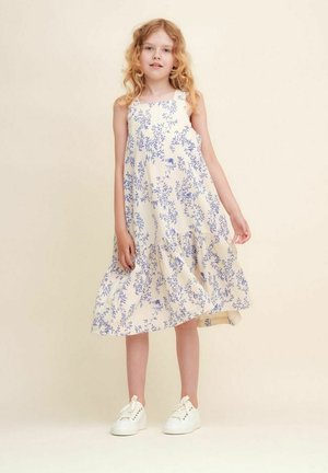 Blomsterkjole i creme med blå mønstre, uden ærmer, ankellang med en lagdelt kant, parret med hvide sneakers.