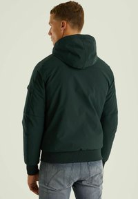 Temno zelena jakna s kapuco in elastičnimi manžetami ter robom. Ima gladko teksturo in minimalističen dizajn, v kombinaciji z svetlo modrimi kavbojkami.