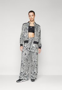 Σαλοπέτα με σχέδιο σε μαύρο και λευκό, που περιλαμβάνει ζακέτα τύπου μπομπέρ με φερμουάρ και παντελόνια με φαρδιά πόδια, και τα δύο με γραφικά σχέδια και επισημάνσεις σε ρίγες.