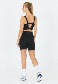 Zwarte sportieve crop top met brede straps en een uitgesneden achterkant, gecombineerd met bijpassende zwarte high-waisted shorts. Witte enkelsokken en schoenen.