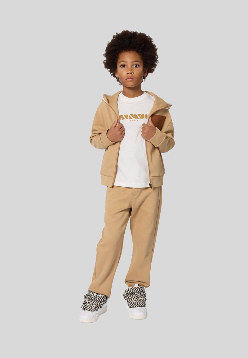 Beżowa bluza z zamkiem, biała koszulka z złotym logo, beżowe dresy i wzorzyste czarno-białe sneakersy. Gładka faktura, luźny krój.