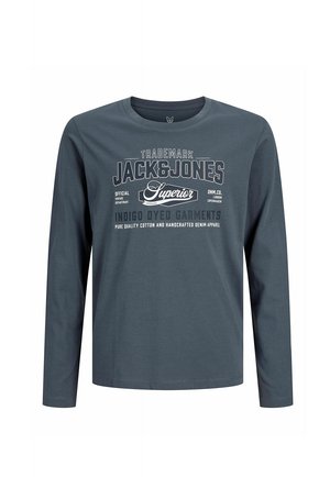 Jongen staat en draagt een grijze Jack & Jones shirt met lange mouwen, zwarte broek en zwart-witte sneakers op een effen lichte achtergrond.