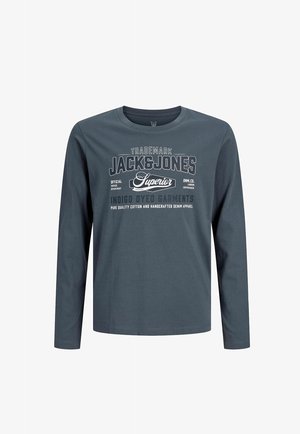 Jongen staat en draagt een grijze Jack & Jones shirt met lange mouwen, zwarte broek en zwart-witte sneakers op een effen lichte achtergrond.