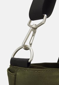 Sac en tissu vert olive avec une sangle noire, équipé d'un clip métallique argenté et d'un anneau, présentant une surface texturée et des coutures renforcées.