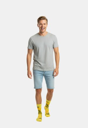 Lachende man in een grijs t-shirt, lichtblauwe denim shorts en felgele sokken met fruitpatronen, lopend naar voren op een effen achtergrond.