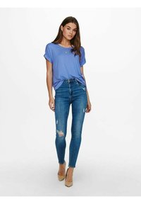 Blå kortermet t-skjorte kombinert med tettsittende slitte blåjeans og beige høye hæler; modellen står med hendene på hoftene mot en ensfarget bakgrunn.