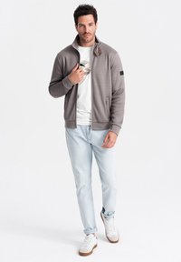 Felpa grigia da uomo con zip, dalla trama a coste, colletto rialzato, due tasche frontali, abbinata a jeans azzurri e sneakers bianche.