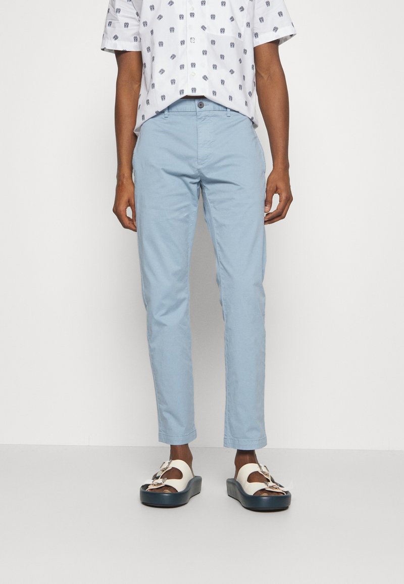 Homme portant un pantalon slim ajusté bleu clair, une chemise à manches courtes blanche à motifs, et des sandales blanches avec des semelles épaisses noires et des boucles.
