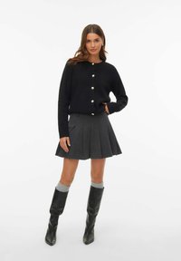 Kvinna som bär en svart knappad cardigan, grå veckad kjol, gråa strumpor och svarta knähöga stövlar, stående med ena handen på höften.