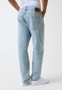 Jean en denim bleu clair à coupe décontractée, avec deux poches arrière et une étiquette en cuir marron à la ceinture. Des baskets blanches sont visibles.