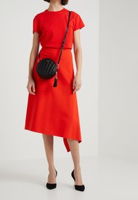 Donna che indossa una gonna rossa asimmetrica e un top rosso a maniche corte, una borsa a tracolla nera rotonda e scarpe con tacco nero.