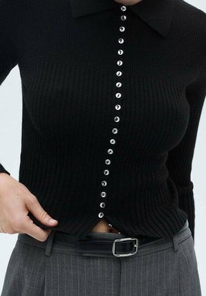 Woman fastening black ribbed button-up top with shiny circular buttons, paired with gray pinstripe trousers and black belt.