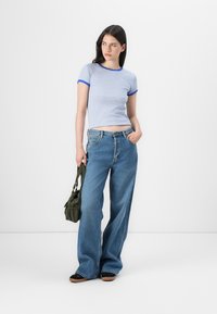 Giovane donna in una camicia a righe bianco e blu e jeans a gamba larga blu, con una piccola borsa verde, si trova di fronte a uno sfondo bianco uniforme.