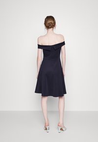 Vestido azul-marinho de ombro à mostra, com corpete ajustado e saia rodada, apresentando um fecho zíper nas costas e um detalhe de colar em camadas.