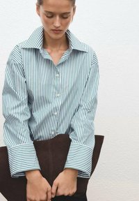 Striped shirt in blue-green and white, button-up design, wide collar, relaxed fit, paired with a brown leather tote, hands holding the bag.