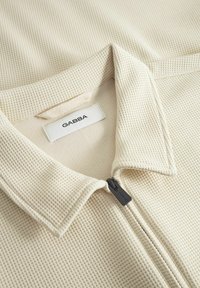 Beige textúrájú zipzáras dzseki gallérral, fekete zipzárhúzóval és fehér címkével, rajta fekete betűkkel "GABBA" felirattal.