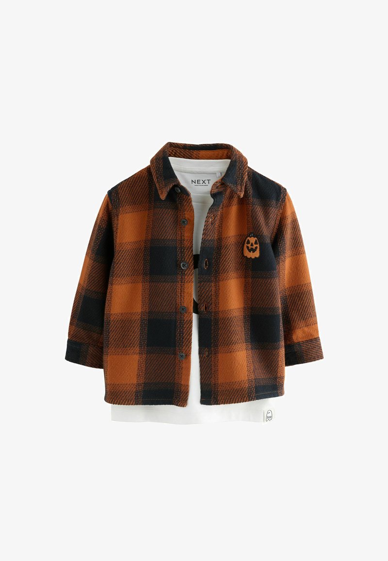 Chemise à carreaux orange et noire boutonnée avec un petit patch visage de citrouille, portée sur une chemise blanche avec un motif fantôme.