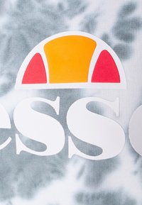 Bílé a barevné logo na šedé batikované látce. Logo má nad tučným bílým textem polosíť v žluté a červené barvě.