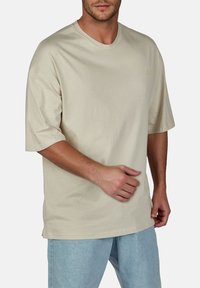Beige katoenen T-shirt met een ronde hals, korte mouwen, comfortabele pasvorm en subtiele merklogo op de voorkant. Gecombineerd met lichtblauwe jeans.