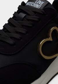 Μαύρο αθλητικό παπούτσι με μαύρα κορδόνια, λευκή σόλα, χρυσή καρδιά σε σχήμα καρδιάς στο πλάι και λογότυπο "LOVE MOSCHINO" στη γλώσσα.