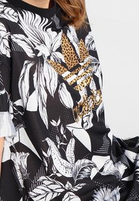 Svart oversize t-shirt med vita blommiga och geometriska mönster, med en leopardmönstrad Adidas-logga i guld på framsidan.