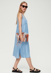 Jasnoniebieska, bezrękawnikowa sukienka dżinsowa o rozszerzanym fasonie, zestawiona z czarnymi sandałami na paski i wielokolorową plecioną torebką.