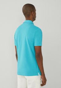 Man i en ljusblå kortärmad polo-shirt och vita byxor, stående vänd mot en enkel ljusgrå bakgrund.