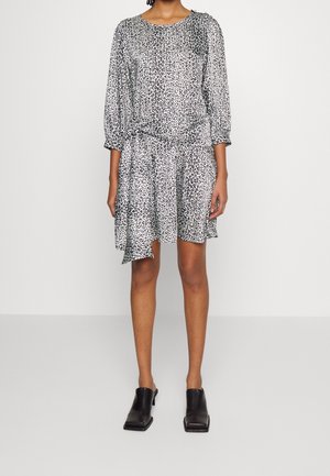 Femme portant une robe à manches longues imprimée léopard noire et blanche avec un ourlet asymétrique, associée à des mules noires à talons.