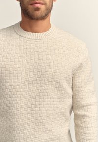 Stickad creamfärgad tröja med ett texturerat kvadratmönster, rund halsringning och ribbad kant. Materialet verkar mjukt och varmt.