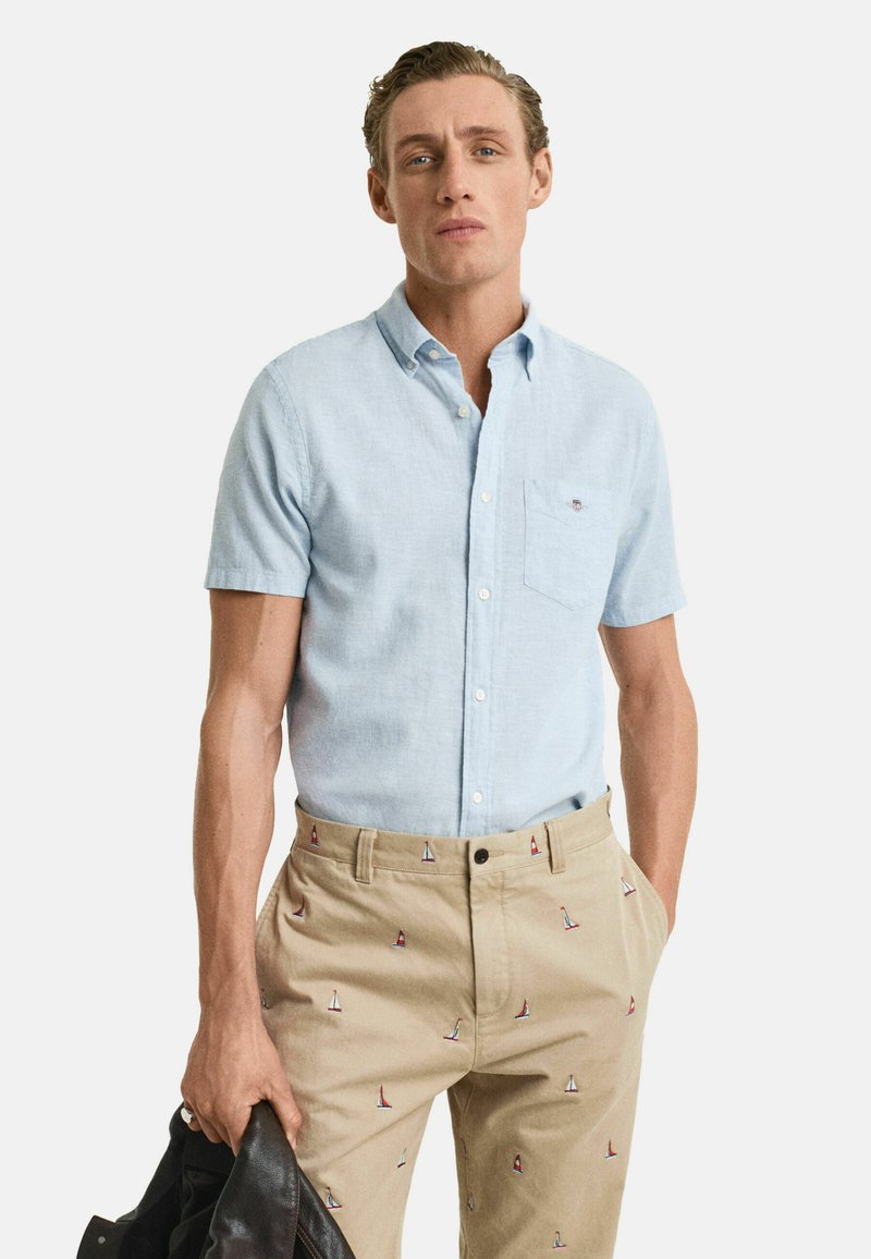 Lichtblauwe kortärmelige button-up shirt met een borstzak, gecombineerd met beige broek met geborduurde zeilbootpatronen. Model houdt een zwarte jas vast.