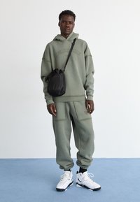Sweat à capuche oversize vert olive avec logo, pantalon baggy assorti et baskets blanches avec des accents noirs. Sac bandoulière noir attaché.