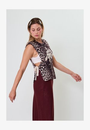 Donna che indossa un gilet senza maniche in maglia con stampa leopardata e laccetti laterali sopra una maglia bianca e una gonna bordeaux, in posa con le braccia tese.