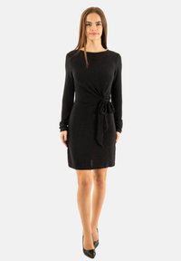 Abito lungo nero a maniche lunghe realizzato in materiale morbido, con una vita aderente e un dettaglio in nodo legato, e orlo a lunghezza ginocchio.