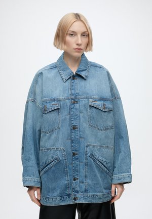 Jeune personne aux cheveux blonds courts portant une veste en denim bleu clair oversize avec une fermeture par boutons et des poches avant.