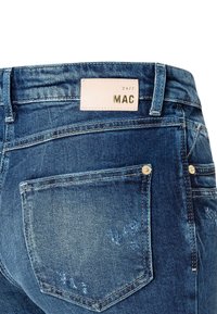 Tmavě modré džíny z denimu s omšelým vzhledem, se koženou nášivkou s nápisem "MAC" a kovovými nýty na zadních kapsách.