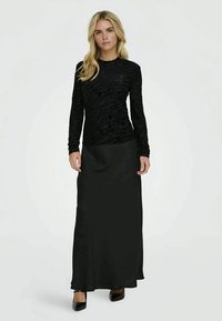 Blonde woman standing wearing a black textured long-sleeve top, black ankle-length skirt, and black pointed heels on a white background.